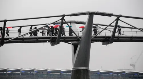This urban architecture photograph captures the Millennium Bridge in London, United Kingdom, during a rainy afternoon in winter. The main subject of the image is the Millennium Bridge, which is seen carrying numerous pedestrians, many of whom are holding umbrellas as they cross. The scene highlights the distinctive design of the bridge and its role as a key architectural landmark in central London, with the overcast sky and muted light typical of winter afternoons. The backdrop includes modern city infrastructure, further emphasizing the urban setting of this London bridge.